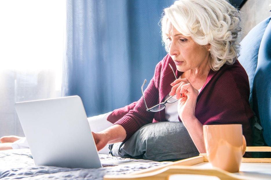 Woman with gray hair in a burgundy cardigan reclining in front of her laptop while holding glasses in her left hand